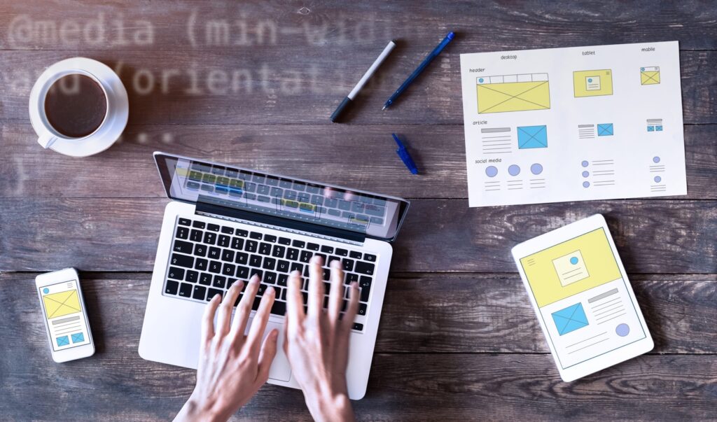 Website wireframes shown on a laptop, smartphone, tablet and paper at a wooden desk.